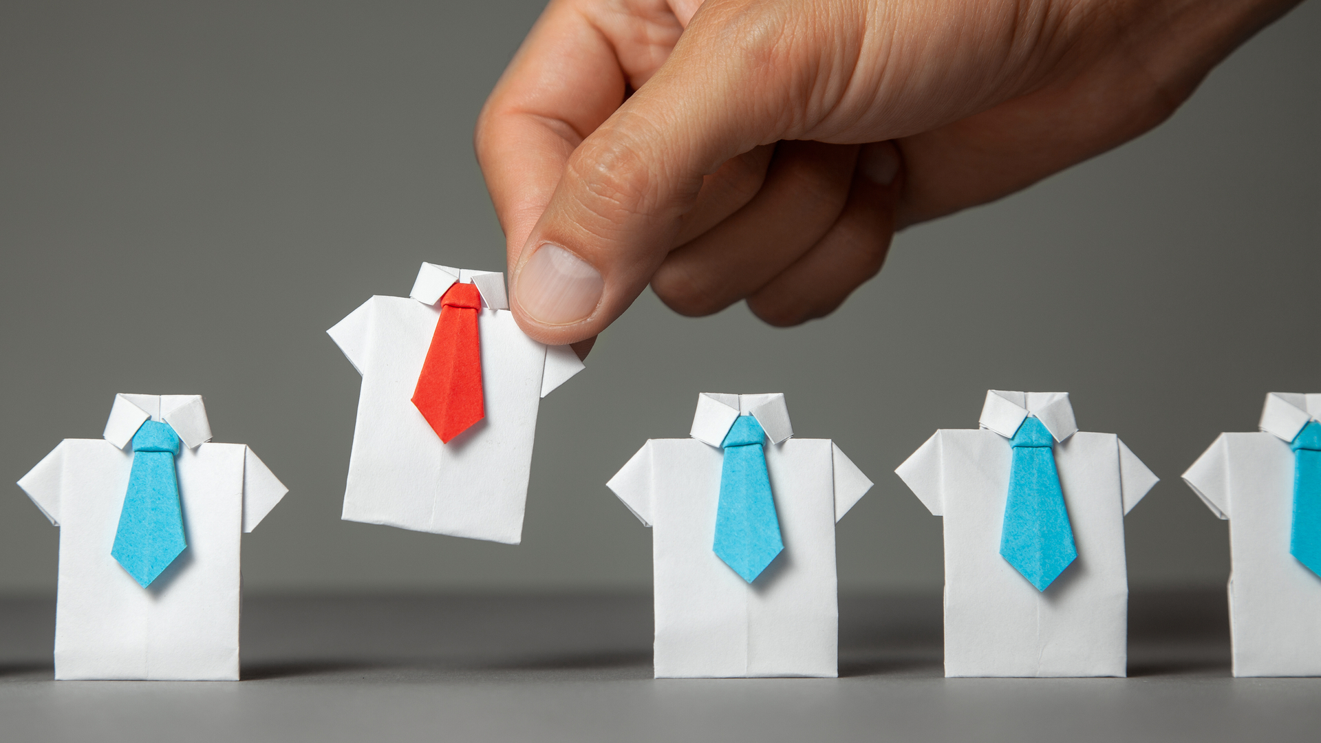 An image of a hand picking a miniature paper shirt with a red tie over other shirts with blue ties. Choosing an adviser in finance