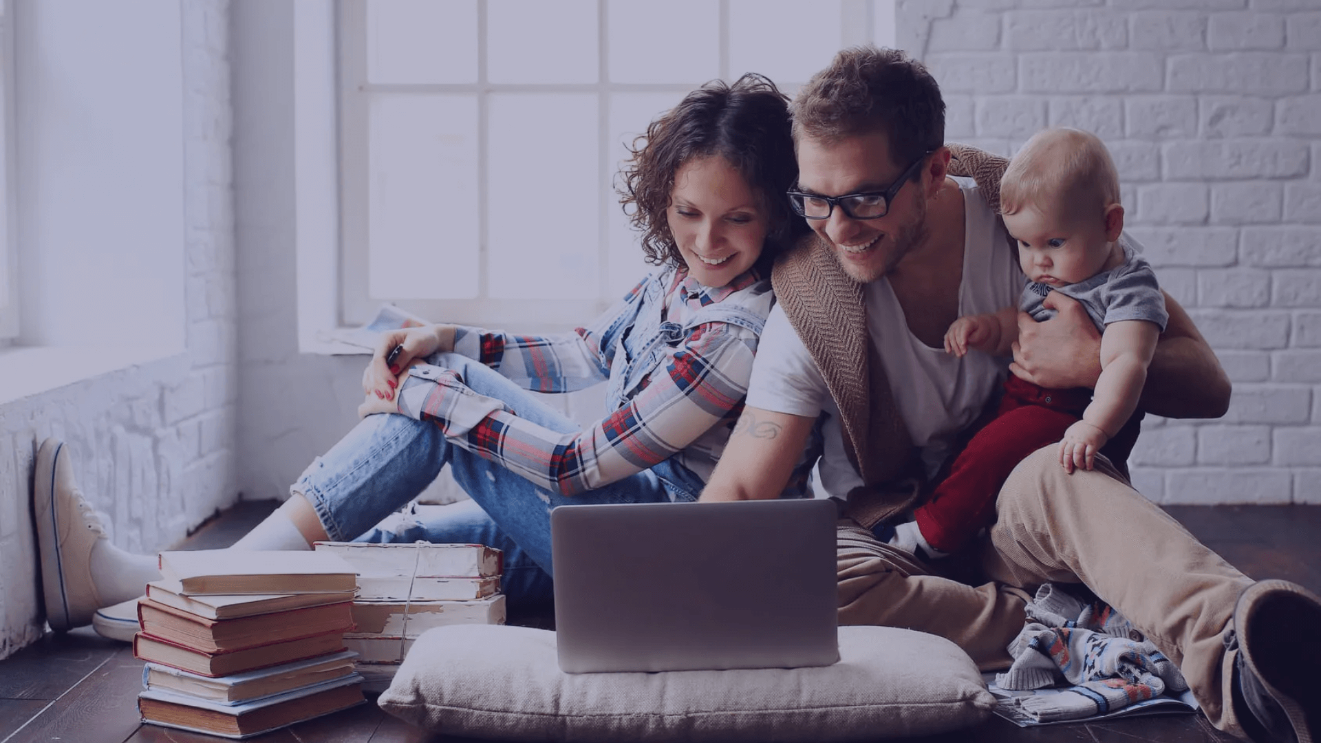 An image of a family. A man, a woman and a baby are looking at the computer with piles of books by them. different life stages and life insurance