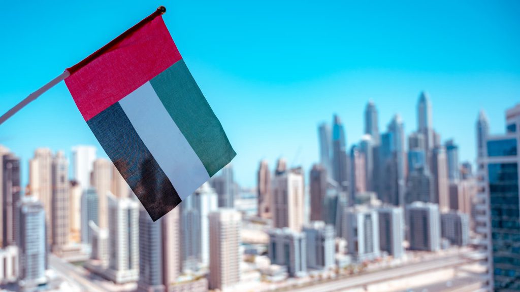 An image of the UAE flag being waved with a sight of a city in the UAE in the background. Guide to moving from the UK to the UAE